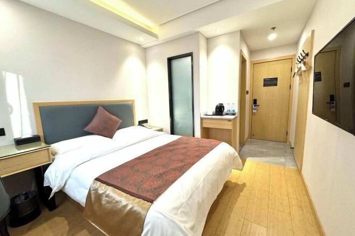 A hotel room with a bed, bedside table, and a desk with a chair. There is a door leading to the bathroom and a mirror on the wall.
