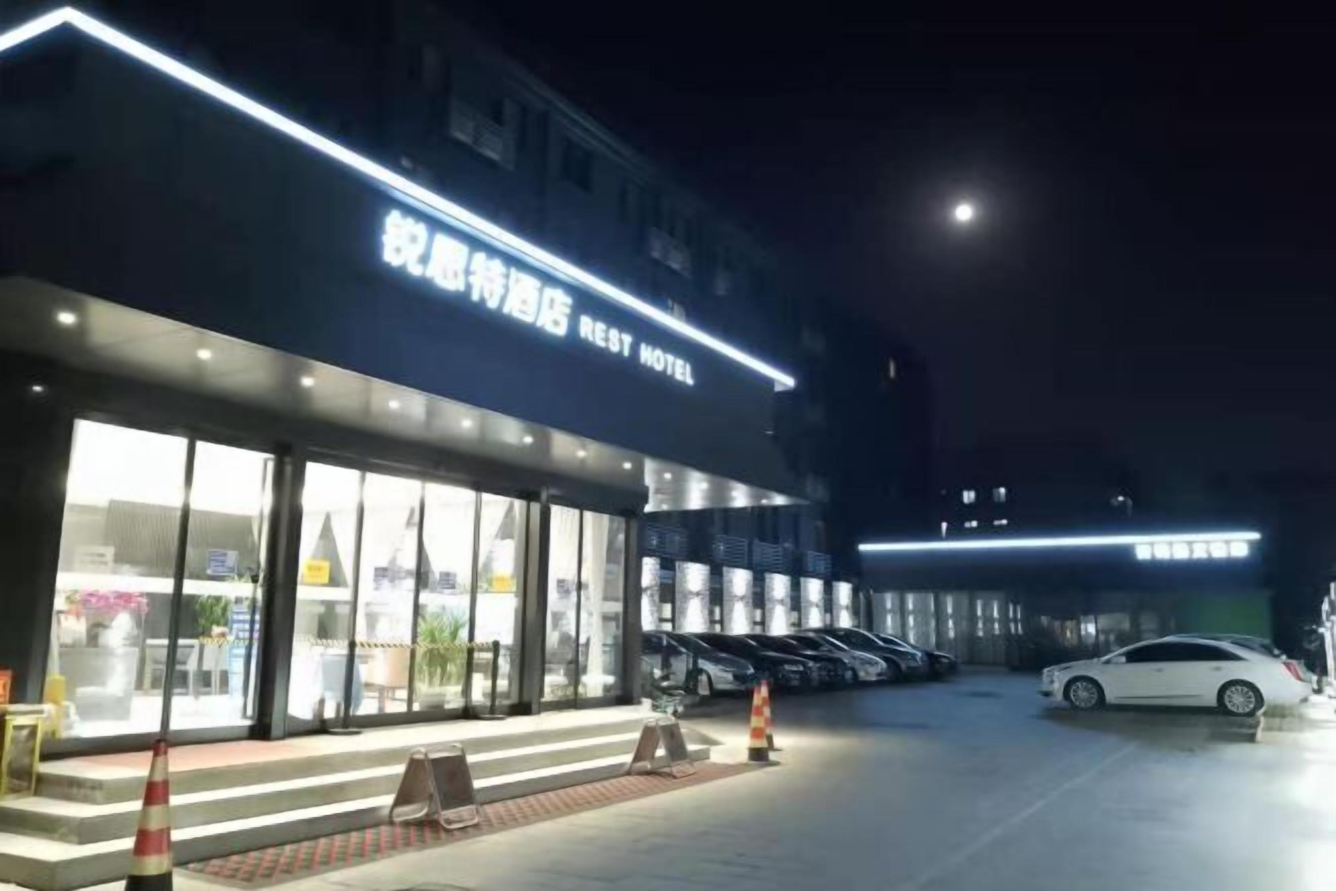 A hotel entrance with a sign reading "Rest Hotel" and a parking area with several cars.