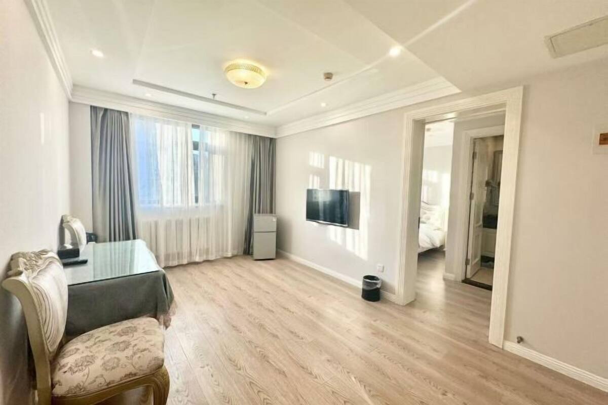 A spacious room with a wooden floor, a dining table with chairs, a television, and a large window with curtains.
