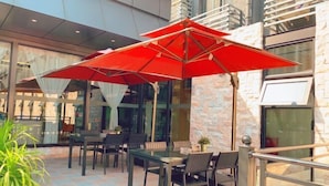 Um pátio externo com mesa e cadeiras de vidro, um guarda-sol vermelho e uma parede de pedra.