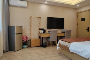 Ett hotellrum med en säng, ett skrivbord med en stol, en tv, ett kylskåp och en liten växt.