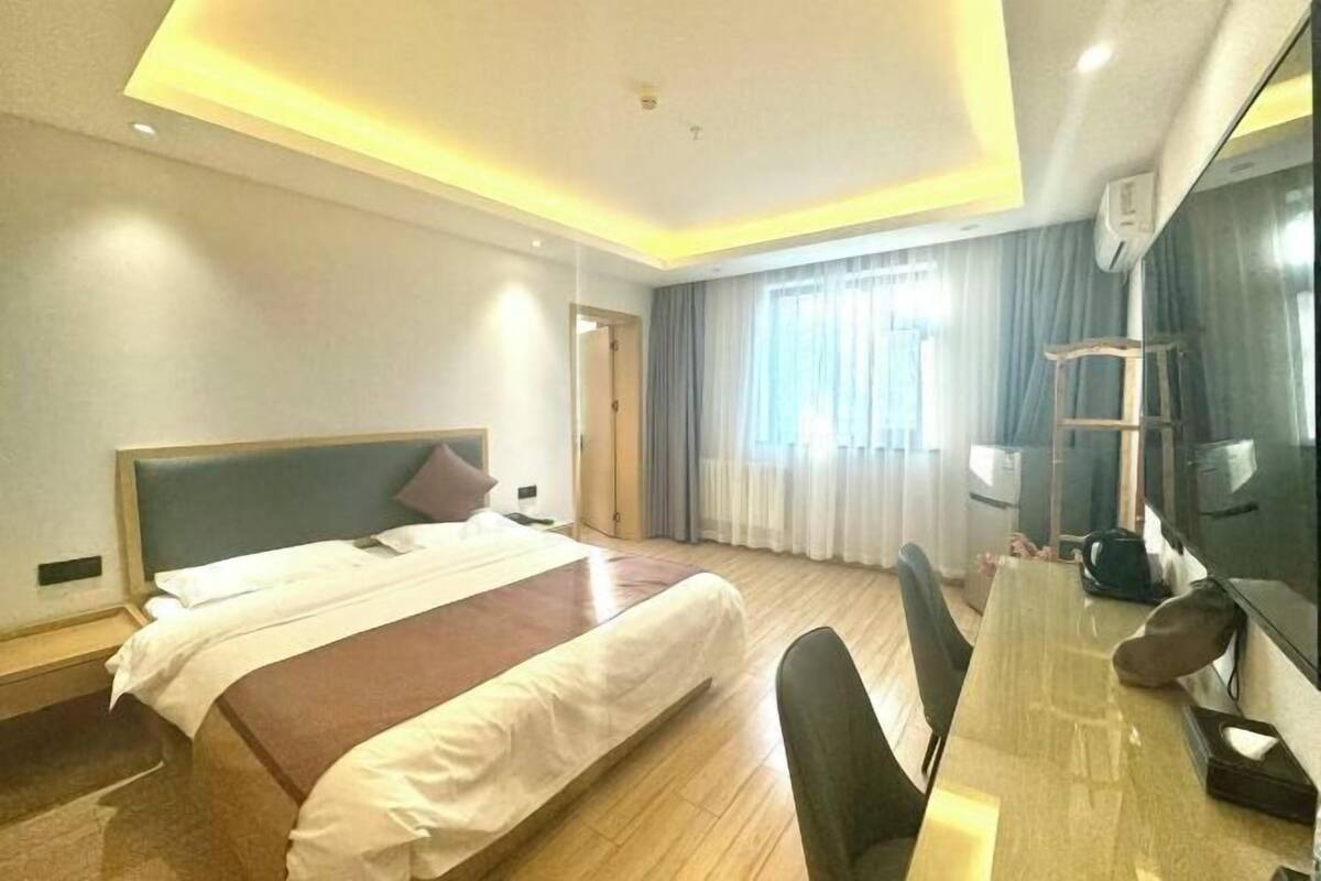 A hotel room with a bed, a desk, and a chair. There is a window with curtains, a mirror, and a ceiling light.