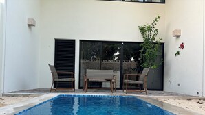 Une piscine, deux fauteuils en osier et une petite table, le tout entouré d’un mur blanc percé d’une porte coulissante et agrémenté d’une plante en pot.