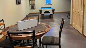 Una habitación con mesa de billar, portaobjetos para billar y un estante para tacos de billar. Hay una mesa con sillas y una ventana con persianas.