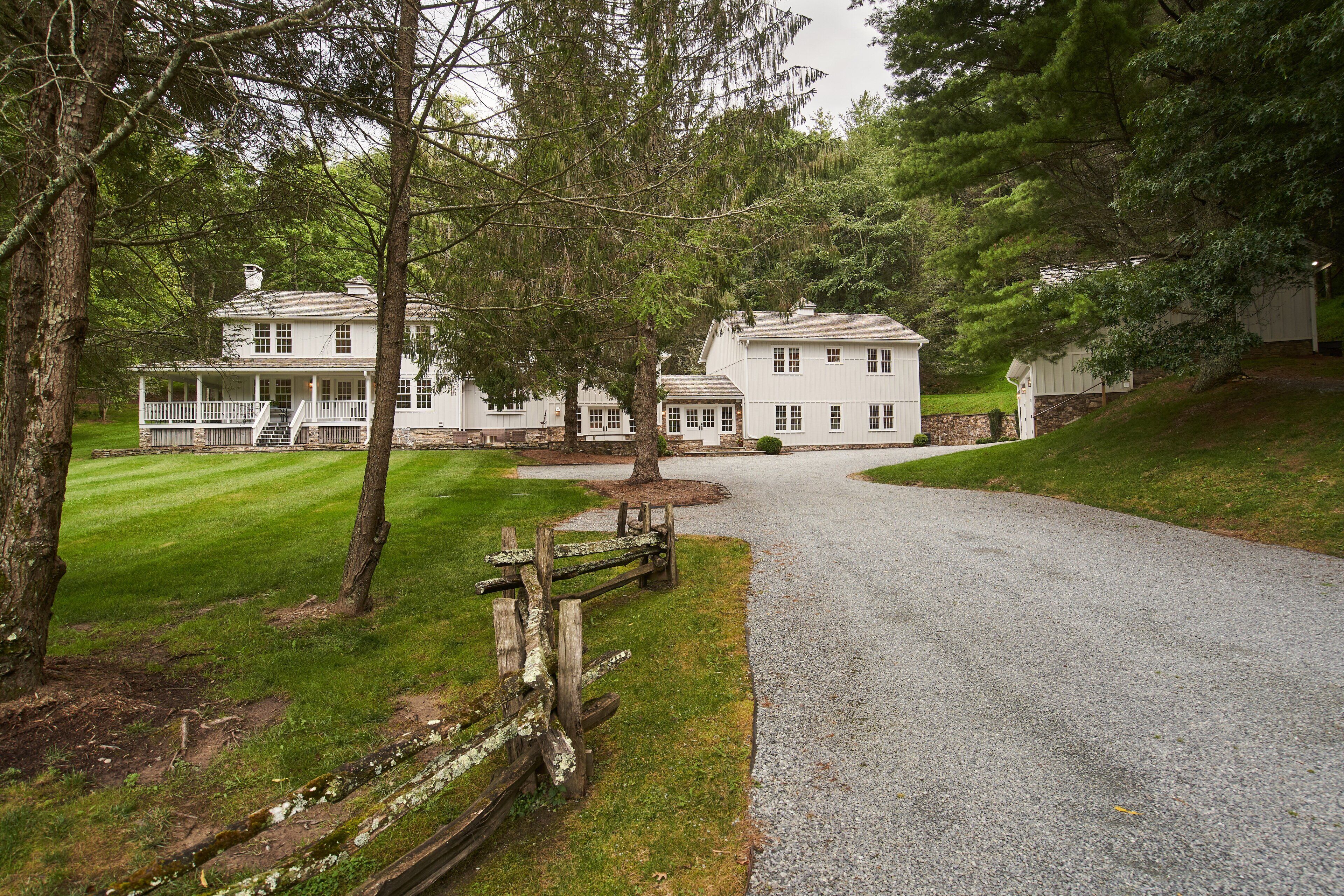 A white two-story house with a porch, surrounded by trees and a gravel driveway.