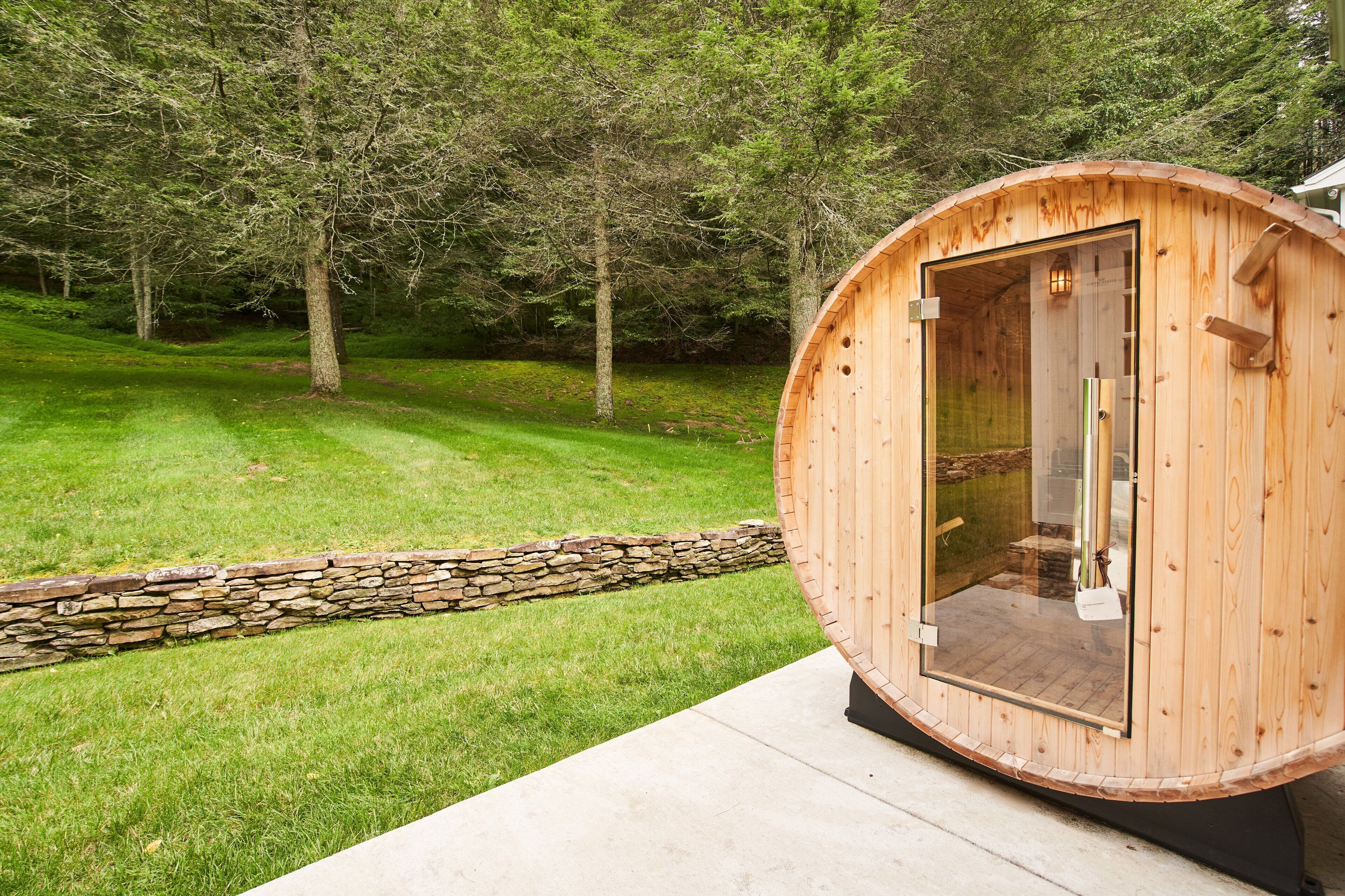 Un sauna en bois doté d’une porte en verre, installé sur une plateforme en béton avec un mur en pierre, dans un espace verdoyant entouré d’arbres.
