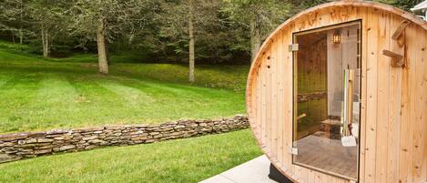 Un sauna en bois doté d’une porte en verre, installé sur une plateforme en béton avec un mur en pierre, dans un espace verdoyant entouré d’arbres.