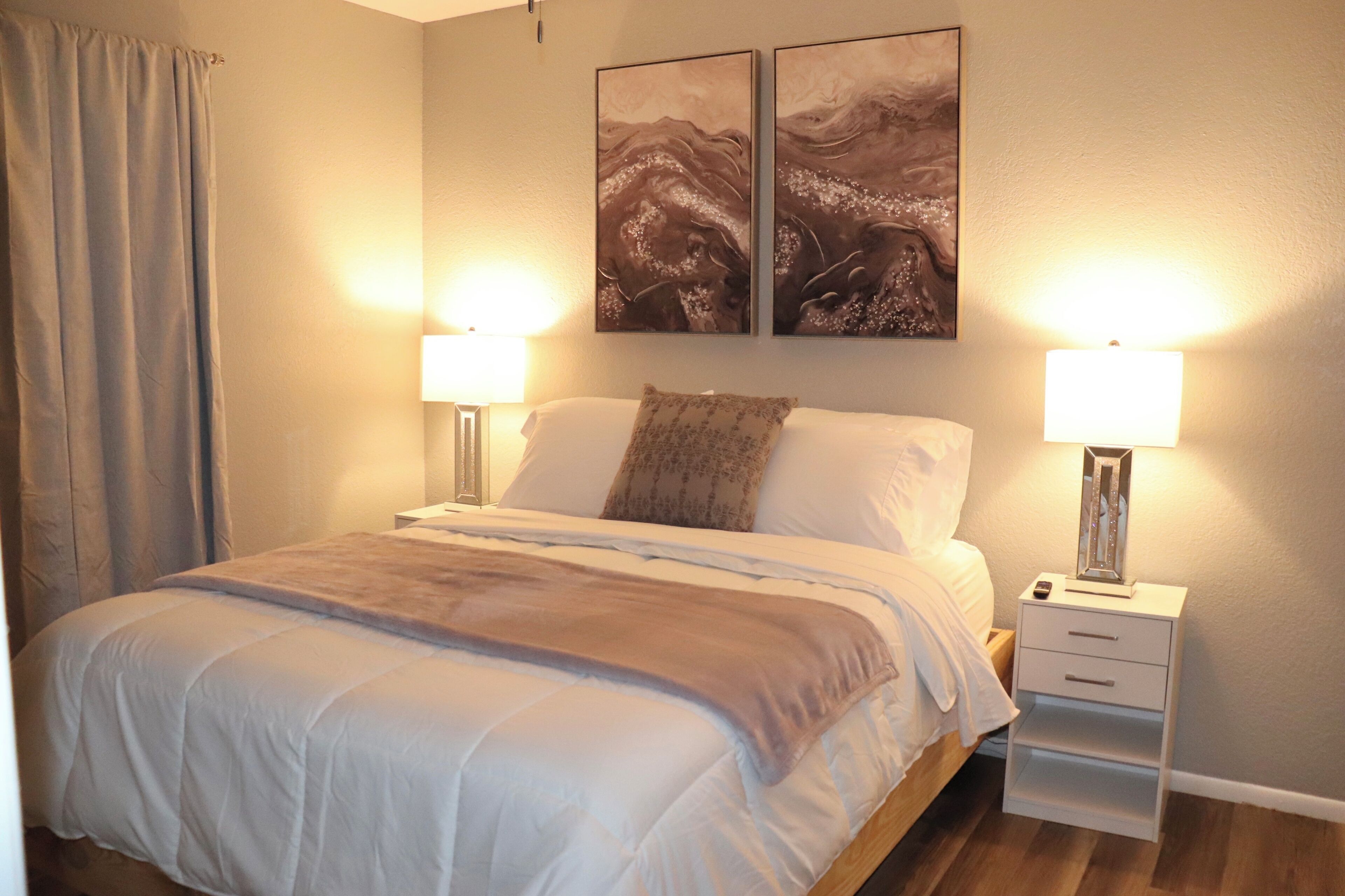 A bedroom with a bed, two bedside lamps, a nightstand, and two wall-mounted paintings.