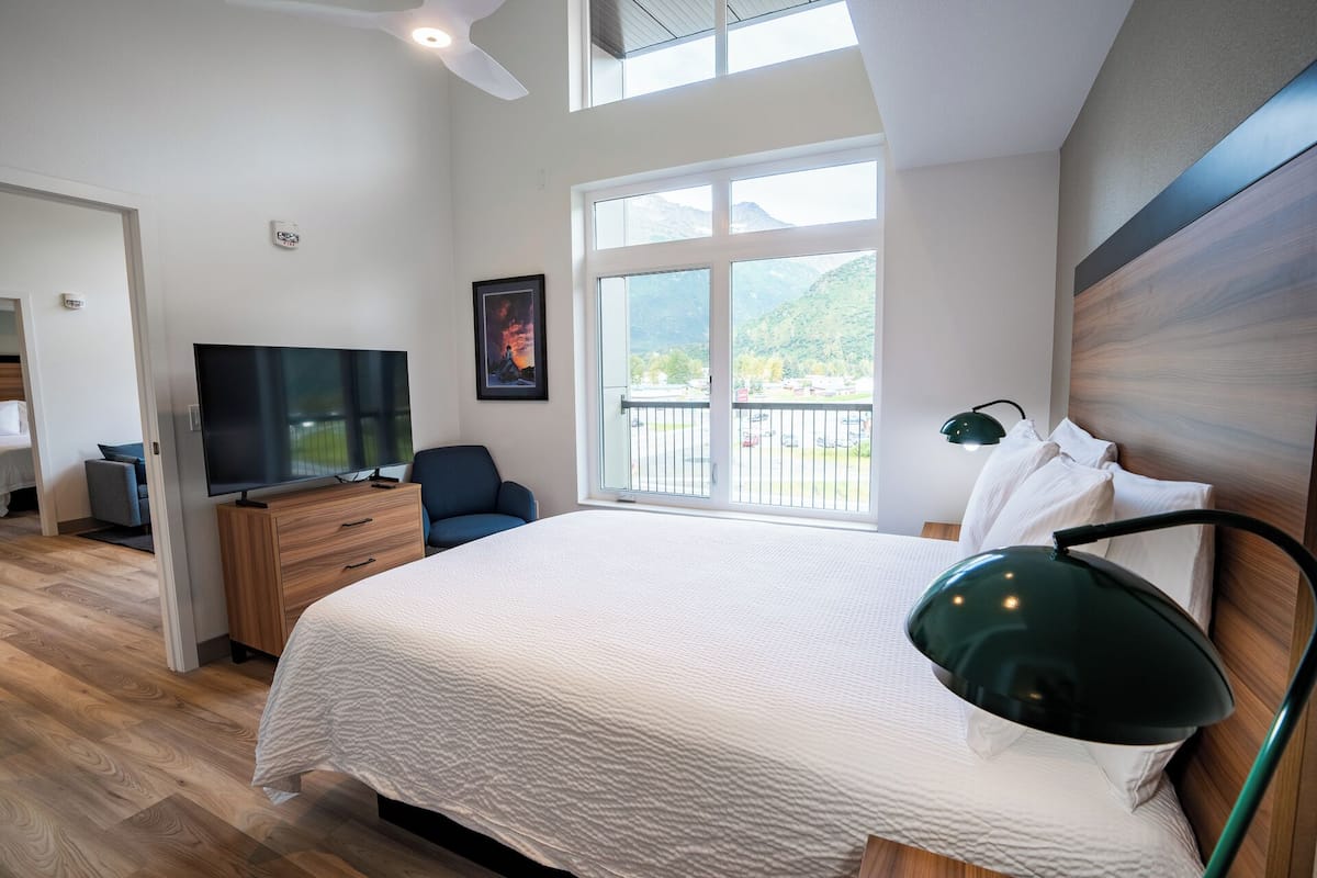 A modern hotel room with a large bed, a TV, a desk, and a balcony with a mountain view.