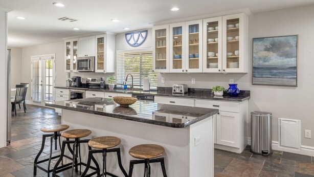 A modern kitchen with white cabinets, a central island with stools, and a large window with blinds.
