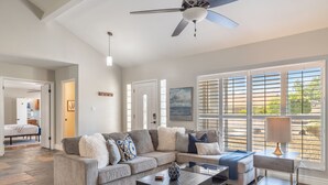 A spacious living room with a sectional sofa, a coffee table, and a side table with a lamp. There are large windows with blinds, a ceiling fan, and a door leading to another room.