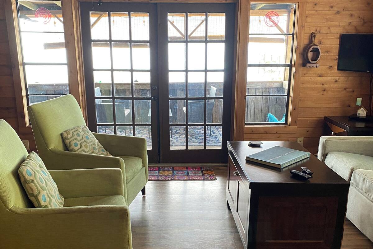 A cozy room with wooden walls and a large sliding glass door leading to a balcony. There is a green armchair with patterned cushions, a wooden coffee table, and a television on a wooden stand.
