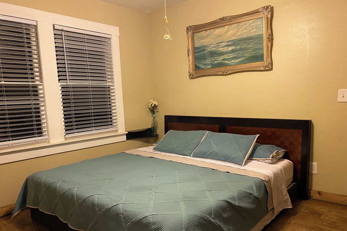 Un dormitorio con cama, ventana con persianas, un cuadro en la pared y un florero con flores.