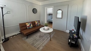 Un salon avec un canapé marron, une petite table ronde ornée d’une plante, un téléviseur et une fenêtre avec un miroir.