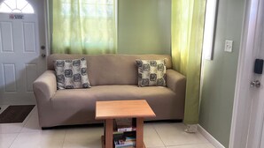 Ein Wohnzimmer mit einem beigefarbenen Sofa, zwei gemusterten Kissen, einem Holztisch und einer Tür mit einem Rauchverbot-Schild.