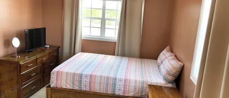 Une chambre à coucher avec un lit, une commode en bois, un téléviseur et une fenêtre avec des rideaux.