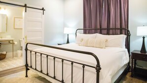Ein Schlafzimmer mit einem Metallbettgestell, weißer Bettwäsche, einem Nachttisch mit Lampe und einem Fenster mit violetten Vorhängen.
