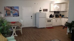 Una cocina compacta con armarios blancos, un refrigerador blanco y un microondas. Hay una mesa de comedor con sillas y un tazón rojo encima. Una planta en maceta está en el suelo, y un cuadro de un auto azul cuelga en la pared.