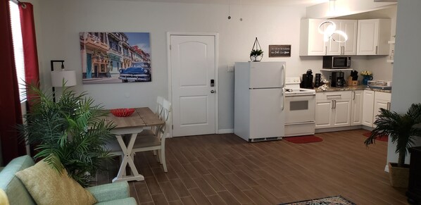 Una cocina compacta con armarios blancos, un refrigerador blanco y un microondas. Hay una mesa de comedor con sillas y un tazón rojo encima. Una planta en maceta está en el suelo, y un cuadro de un auto azul cuelga en la pared.