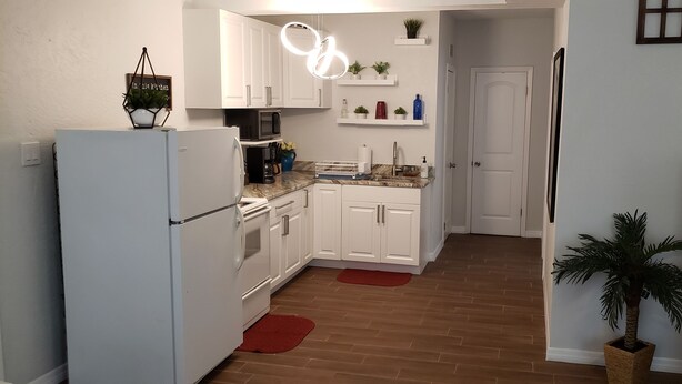 Cocina pequeña con armarios blancos, un microondas y un refrigerador. Hay tapetes rojos en el piso, y se ve una planta en maceta.