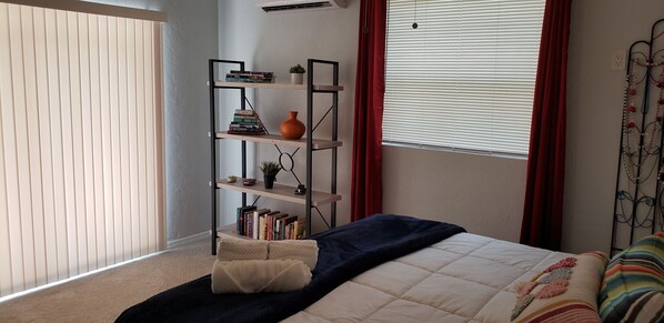 Un dormitorio con cama, una estantería, una ventana con persianas y una cortina roja.