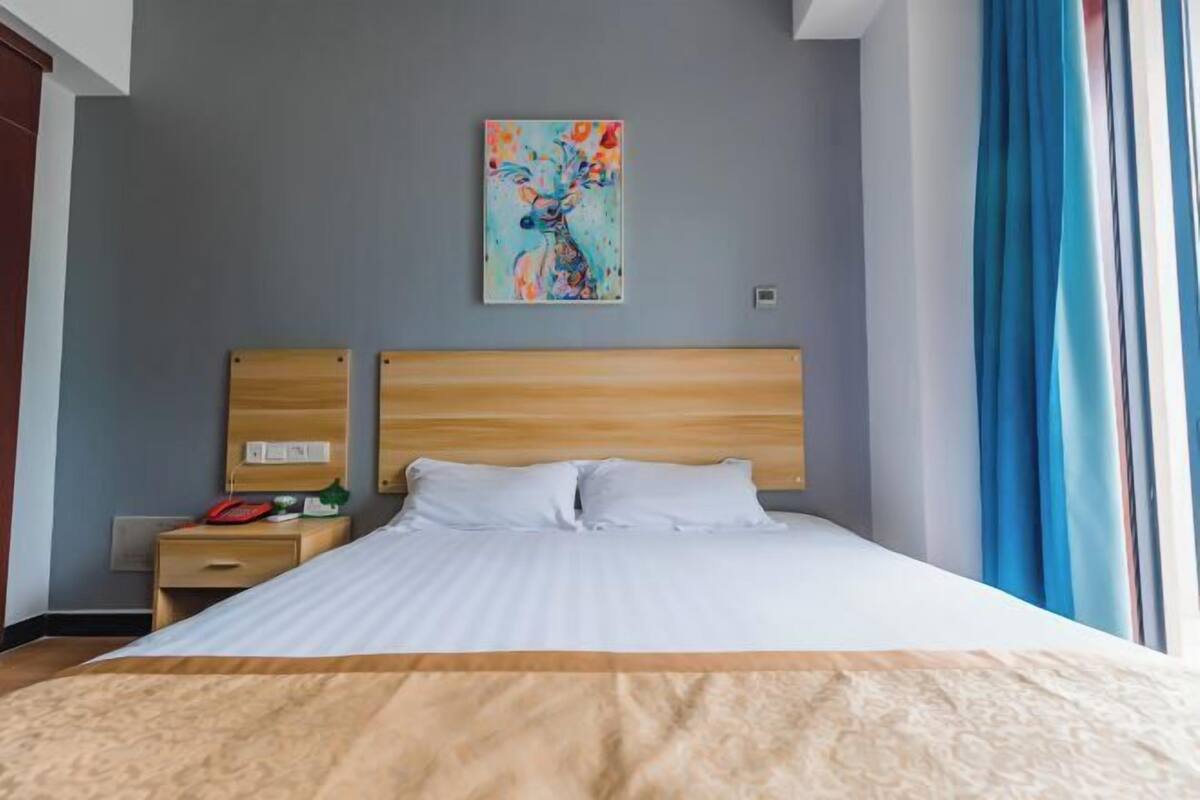 Hotellihuone, jossa on sänky, yöpöytä, taulu seinällä ja ikkuna, jossa on verhot.