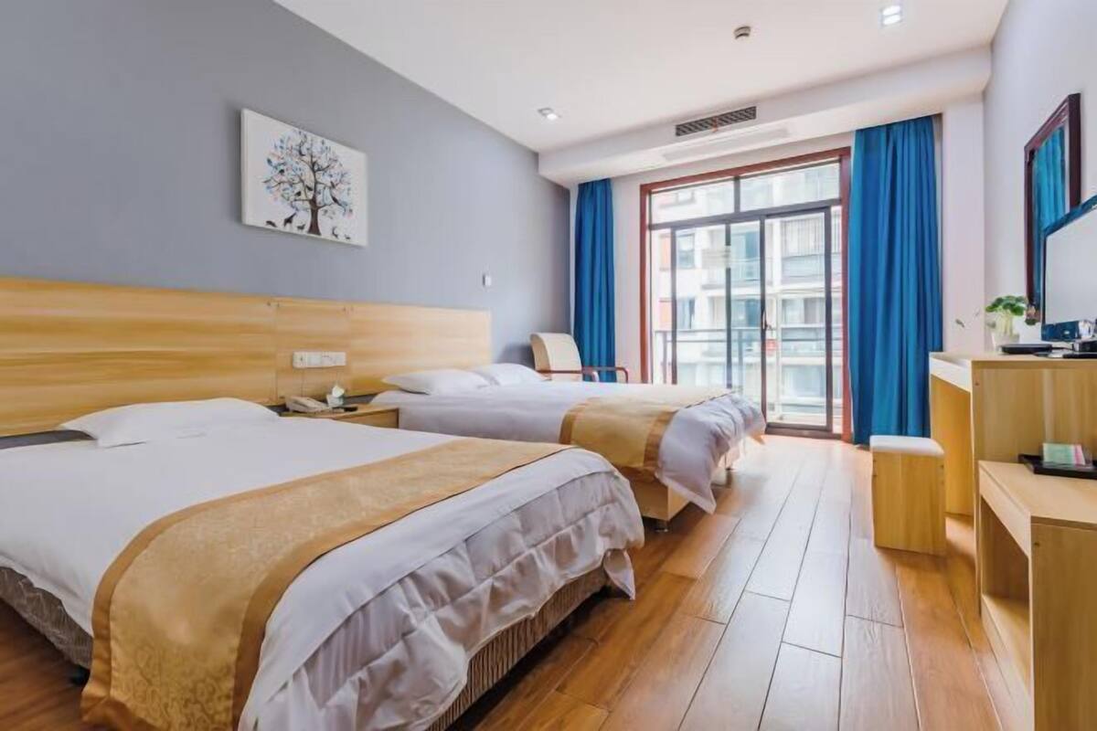 Hotellihuone, jossa on kaksi sänkyä, työpöytä, tuoli, sinisillä verhoilla varustettu ikkuna ja seinällä taulu.