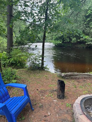 A blue chair by a riverbank with a fire pit nearby. - Secluded Cabin On the Salmon River Peninsula With AC and Heat in Westville NY (Constable)