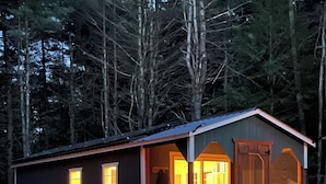 En liten stuga med en veranda, ett fordon parkerat framför och en skog i bakgrunden.