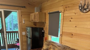 En stuga med en soffa, ett kylskåp, en klocka och ett fönster med utsikt över en terrass.