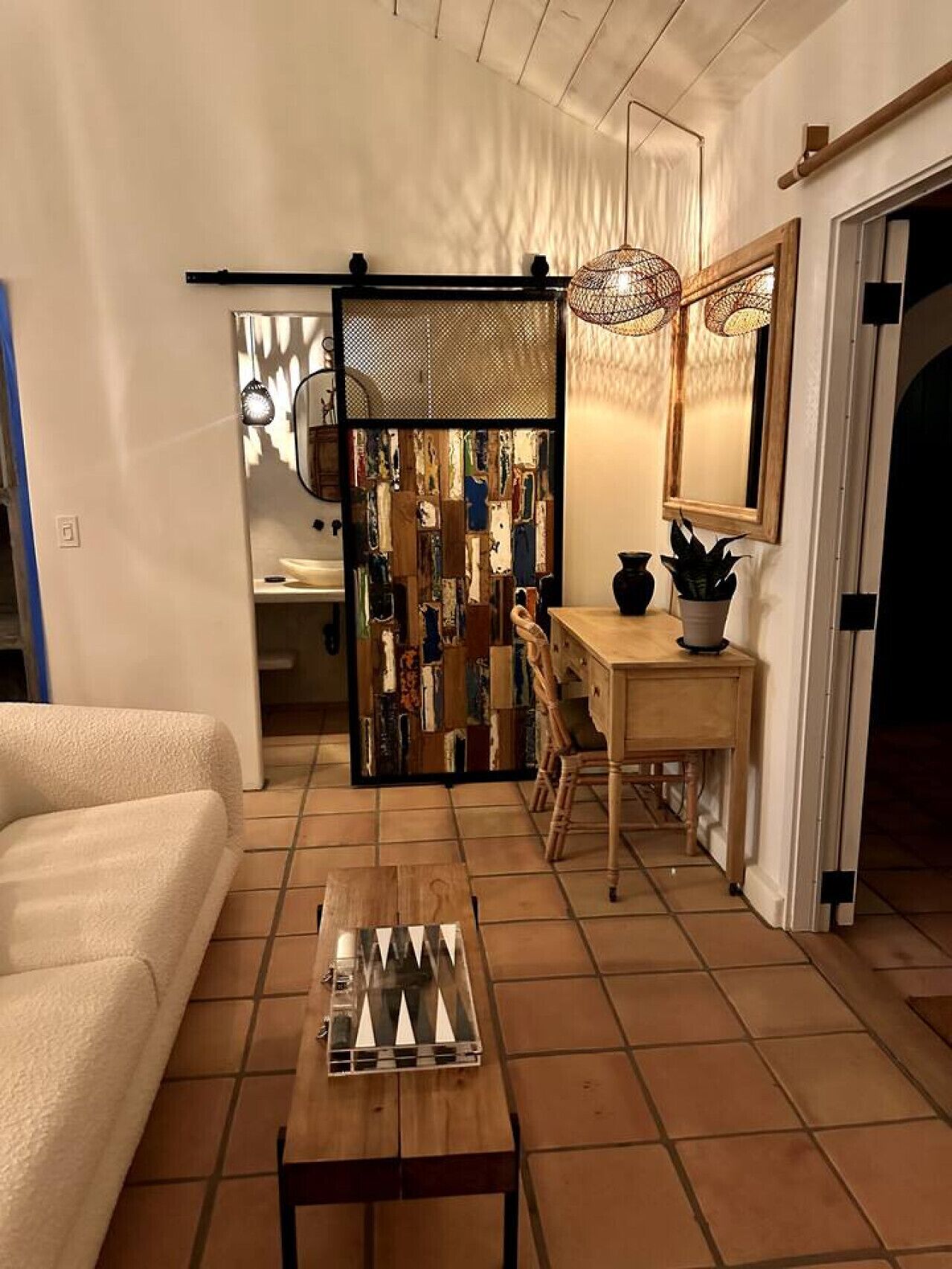 Un salon chaleureux avec une table basse en bois, un canapé, un bureau avec une chaise, une bibliothèque et un grand miroir.