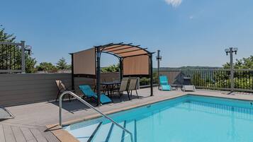 A swimming pool with a wooden deck, lounge chairs, a covered seating area, and a view of the landscape.