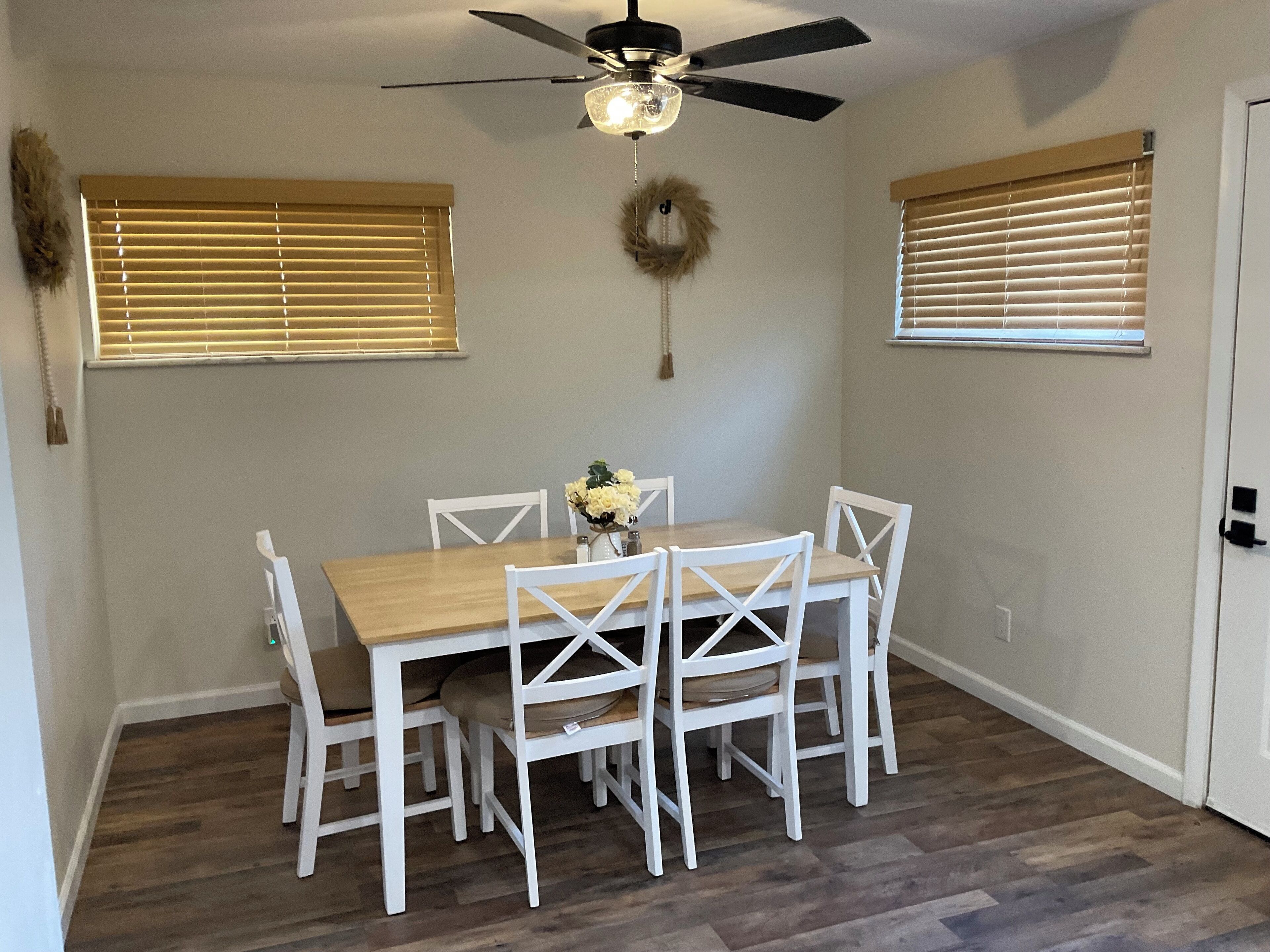 Un coin repas avec une table en bois et des chaises blanches, un ventilateur de plafond et deux fenêtres équipées de stores.