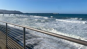 Vista costera con un muelle que se adentra en el mar, una baranda de metal en primer plano y un cielo despejado.