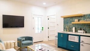 Un salon moderne doté d’un téléviseur à écran plat, d’un fauteuil bleu, d’une table basse en verre et d’un coin cuisine avec des meubles de couleur turquoise et un four à micro-ondes.