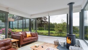 A modern living room with a large window, a fireplace, leather sofas, a coffee table, and a wicker basket. - Pembroke Cottage -  A short stroll from Cartmel Village (Cartmel)