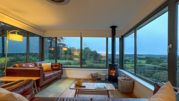 A modern living room with a leather sofa, a wooden coffee table, and a fireplace. Large windows offer a view of the countryside. - Pembroke Cottage -  A short stroll from Cartmel Village (Cartmel)