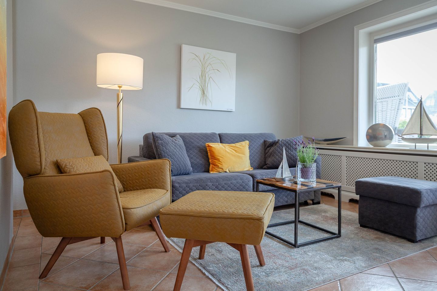 Een moderne woonkamer met een bank, fauteuil, voetenbankje, vloerlamp en een potplant.