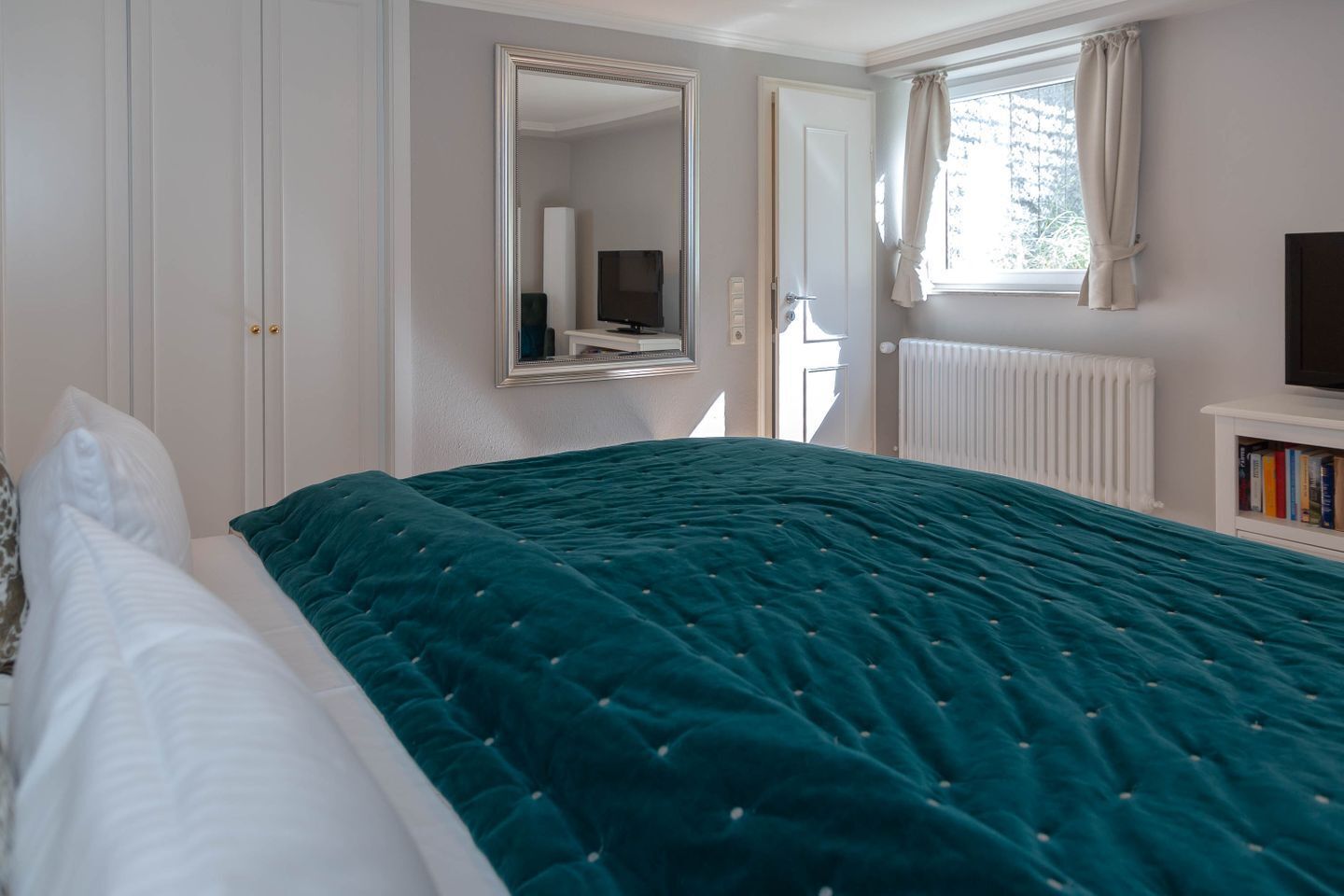 Un dormitorio con cama, espejo, televisor, ventana con cortinas y radiador.