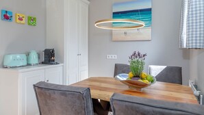 Un comedor con una mesa de madera, sillas, un cuenco de frutas y hierbas, una máquina de café y un cuadro de una playa.