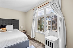 Cozy retreat with a comfortable bed, bedside table, and large window offering a view of the surrounding buildings.