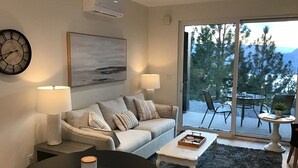 Een moderne woonkamer met een bank, twee bijzettafeltjes, een salontafel en een groot raam met uitzicht op bomen en bergen.