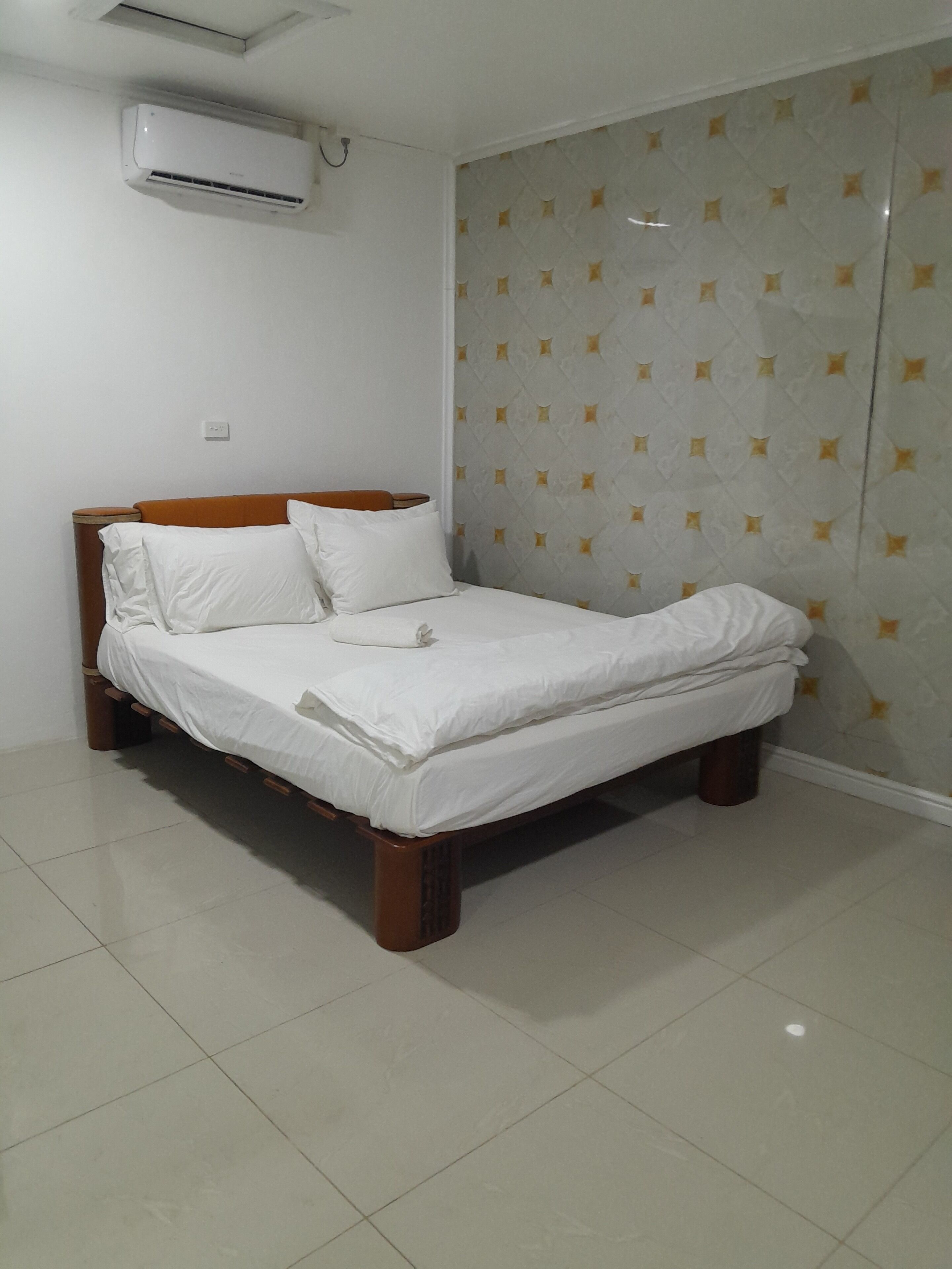 A neatly made bed with white linens, a wooden headboard, and a wall-mounted air conditioner.
