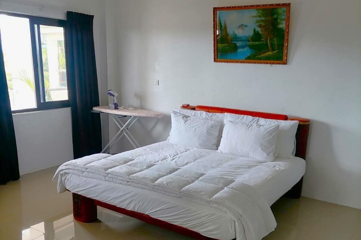A neatly made bed with white bedding in a room with a window, a painting on the wall, and a small ironing board.