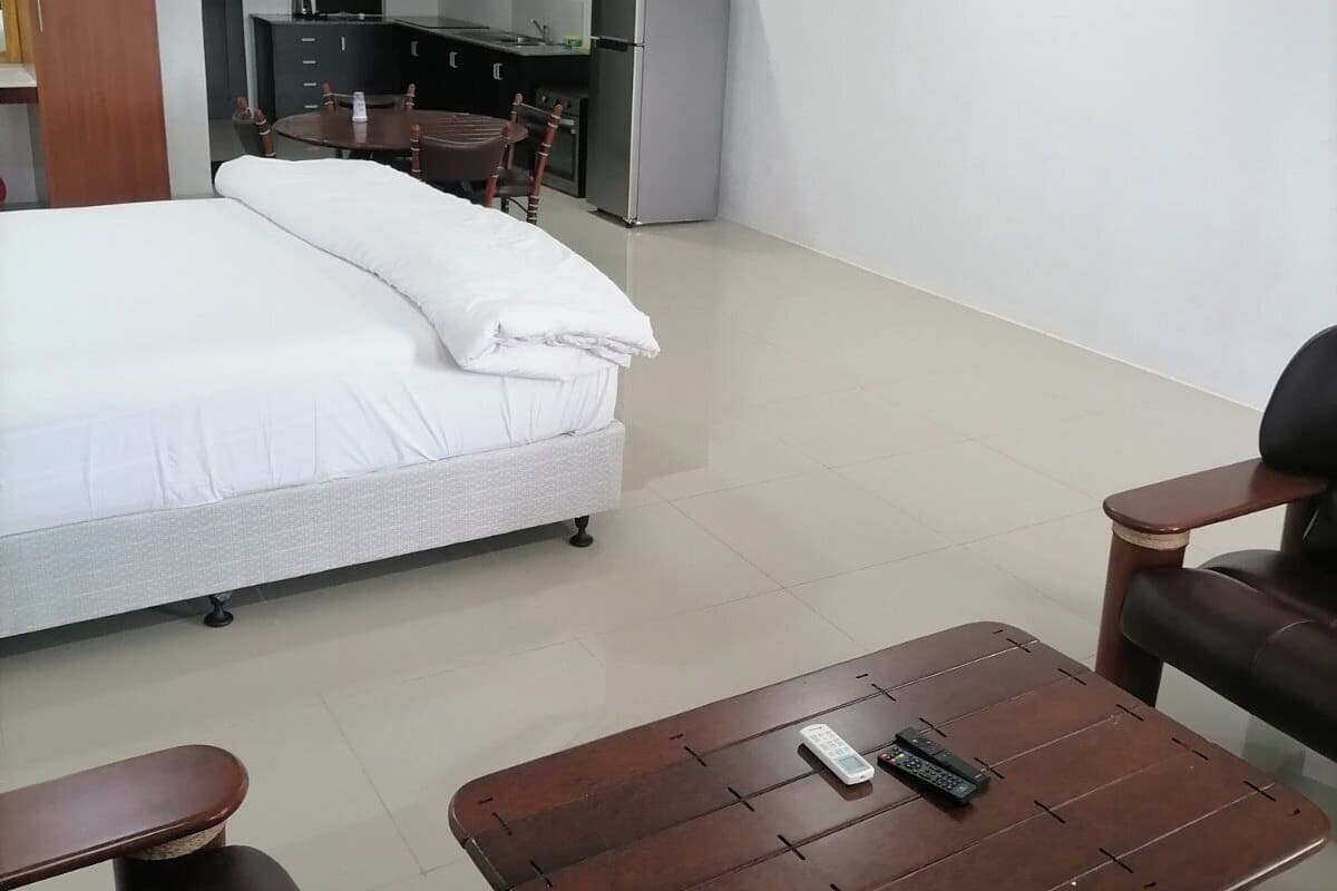 A modern hotel room with a large bed, a wooden coffee table, a leather sofa, a kitchen area with a refrigerator, and a flat-screen TV.
