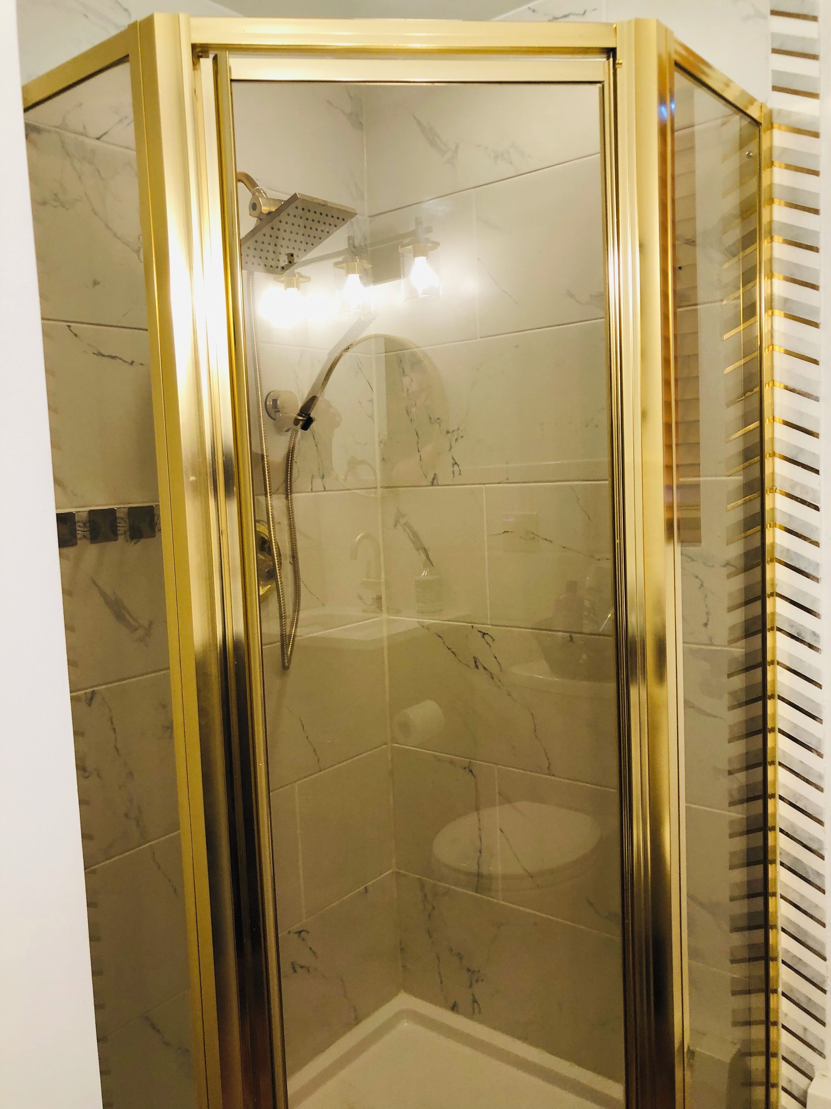A corner shower with a glass door and gold frame, marble tiles, and a handheld showerhead.
