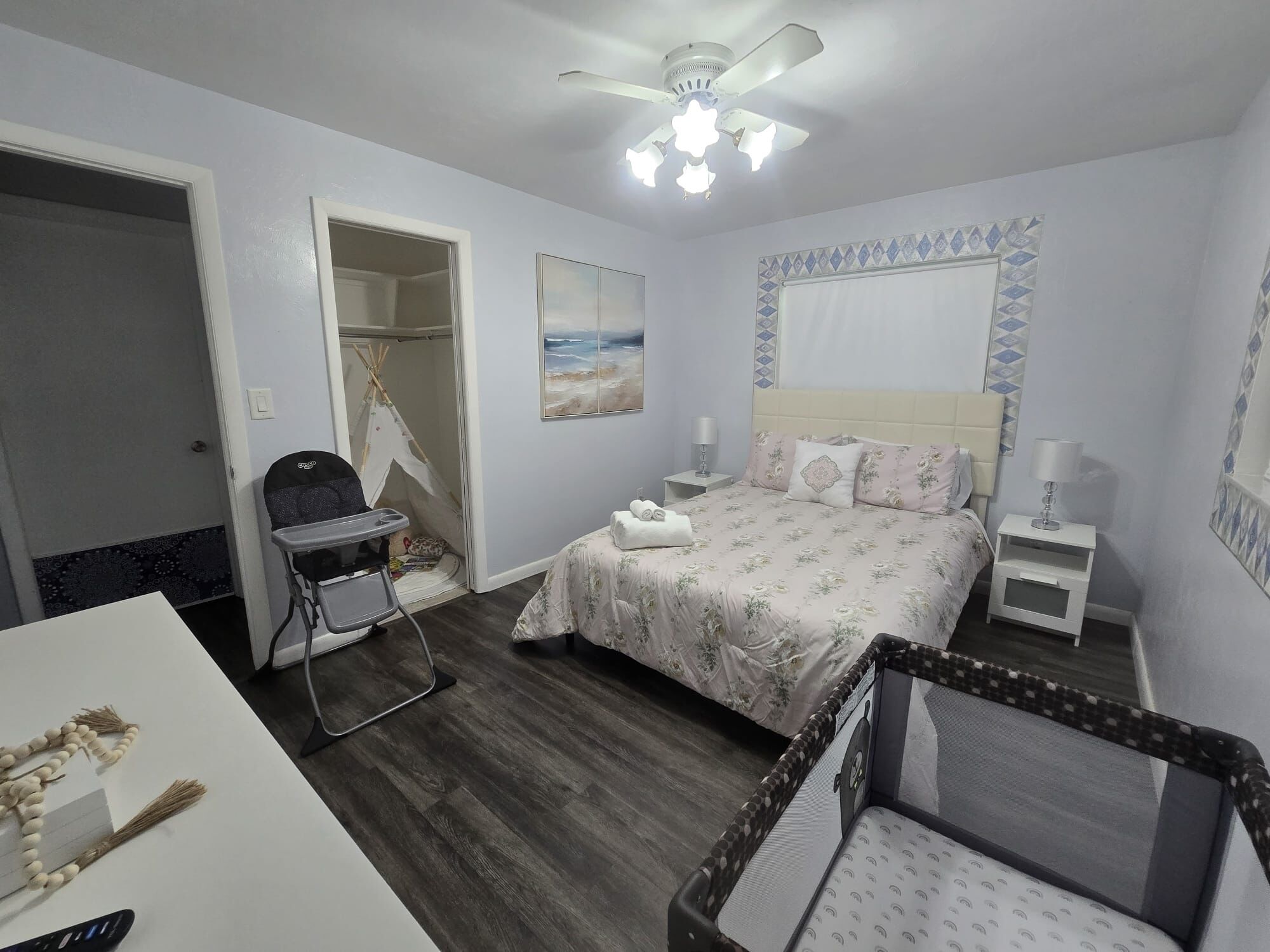 Un dormitorio con cama, mesitas de noche, ventilador de techo, ventana con vistas y puerta que da a otra habitación.