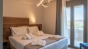 A modern hotel room with a wooden headboard, a large bed with white linens, multiple rolled towels, a knitted throw, and a balcony with a scenic view.
