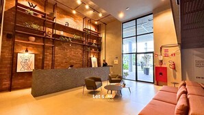 A reception area with a wooden reception desk, two armchairs, a sofa, and a large window with a view of the street. - AX Way Tambaú Suítes (João Pessoa)