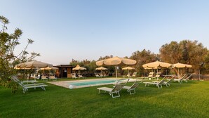 Zona de piscina amb gandules i ombrel·les, un edifici petit i arbres al fons.